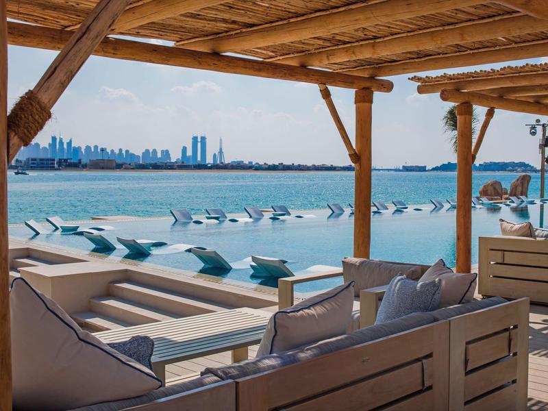 Blick von Strandbalkon mit Holzdach auf Infinity-Pool und Stadtblick am Meer bei klarem Himmel.