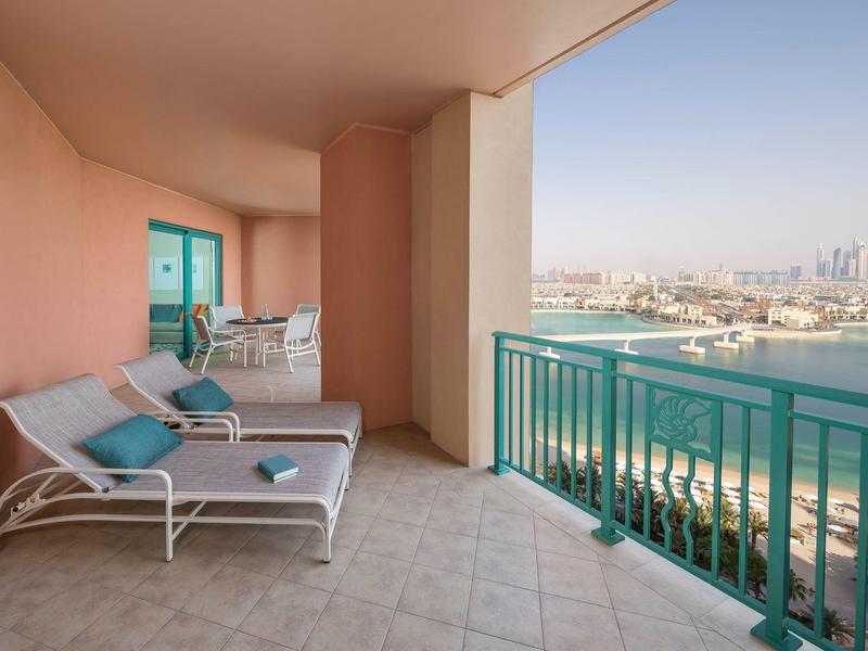 Balkon mit Liegestühlen und Tisch, Blick auf Wasser und entfernte Stadt bei klarem Himmel.