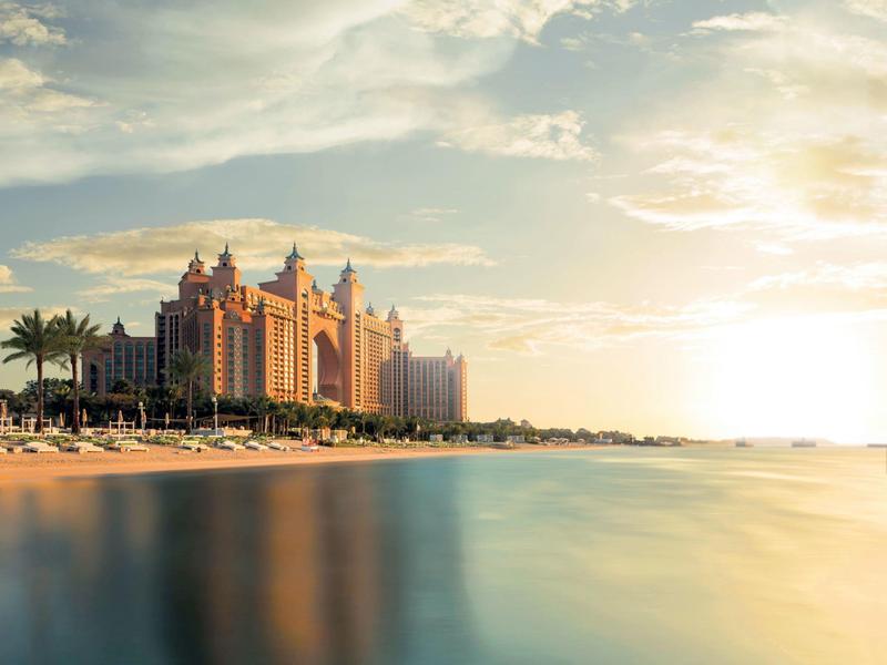 Luxuriöses Hotel am Strand mit Palmen und ruhigem Meer unter hellem Himmel bei Sonnenuntergang.