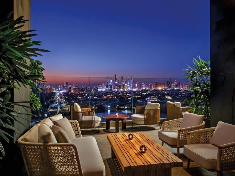 Gemütliche Dachterrasse mit Korbmöbeln, Holztisch und Stadtblick bei Abenddämmerung.