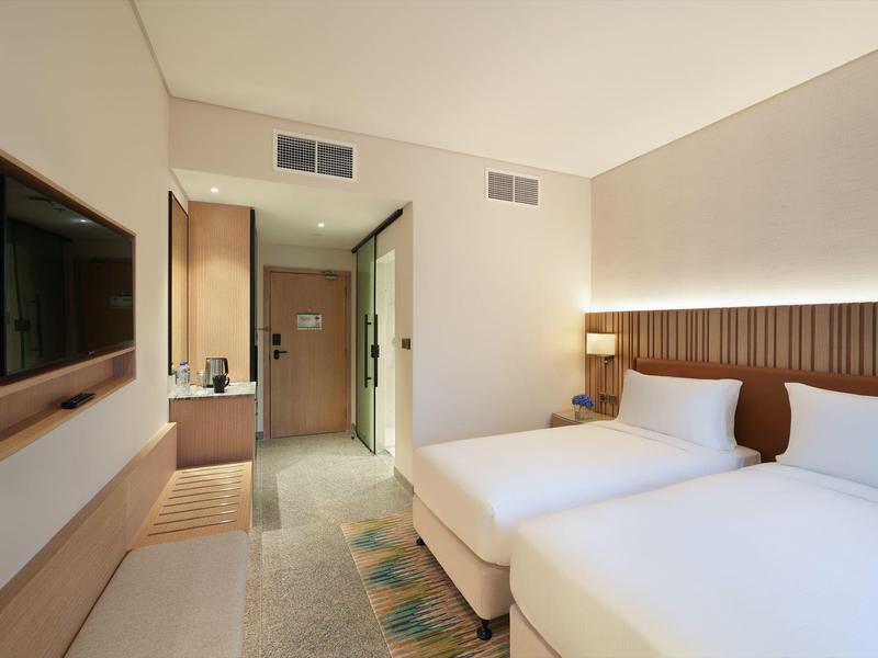 Helles Hotelzimmer mit zwei Einzelbetten, Wand-TV, Holzakzenten und minimalistischem Design.