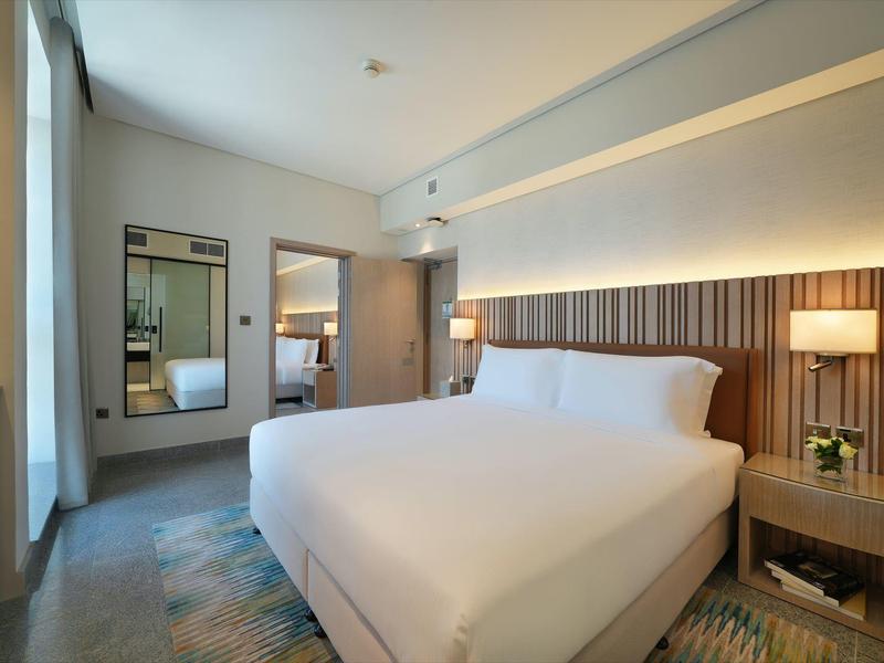 Hell eingerichtetes Hotelzimmer mit großem Bett, Nachttisch, Teppich und großem Spiegel an der Wand.