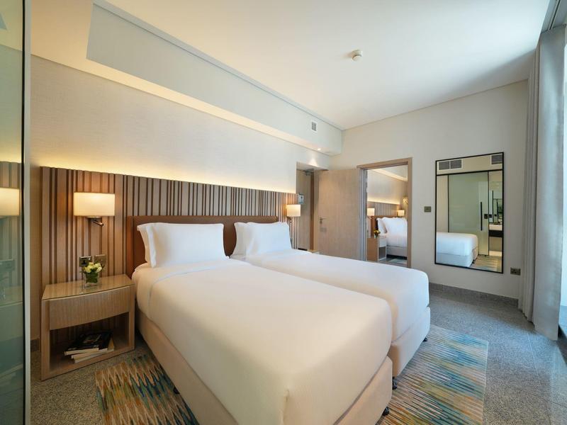 Modernes Hotelzimmer mit zwei Doppelbetten, Nachttischlampen und großem Fenster mit Vorhängen.