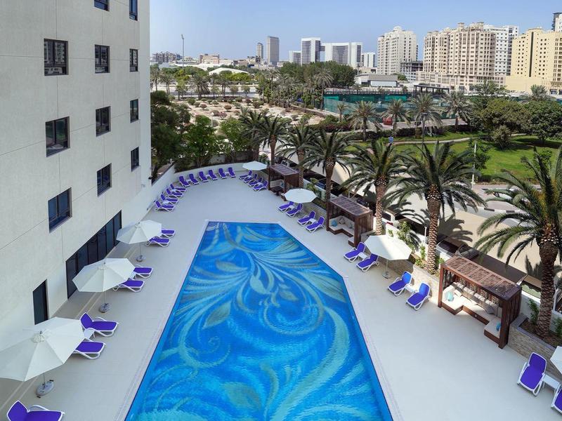 Außenpool mit blauem Wasser, umgeben von weißen Liegestühlen und urbaner Hochhauskulisse.