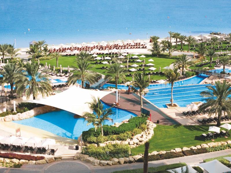 Große Außenpoolanlage mit Sonne, Palmen, Liegen und schattigen Pavillons am hellblauen Himmel.