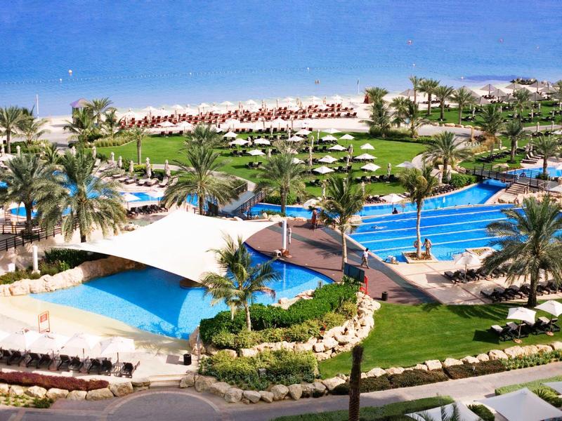 Großer Poolbereich mit blauen Schwimmbecken, grünen Palmen und gepflegten Rasenflächen am Meer.