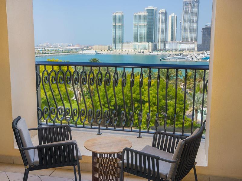 Balkon mit zwei schwarzen Stühlen und kleinem Tisch, Blick auf Wasser, Grünfläche und Wolkenkratzer.