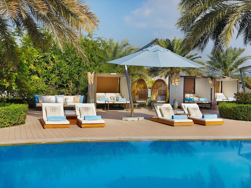 Schwimmbad mit blauer Wasserfläche, Liegen mit blauen Kissen und Sonnenschirmen, Palmen im Hintergrund.