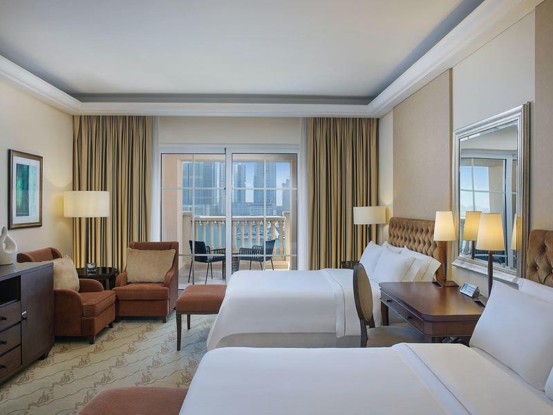 Helles Hotelzimmer mit zwei Betten, Sesseln, Nachttischen, Lampen und Balkon mit Blick nach außen.