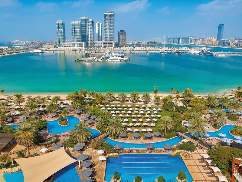 Blick auf Küstenhotel mit Pools, Palmen, grünen Liegen und modernen Hochhäusern im Hintergrund.