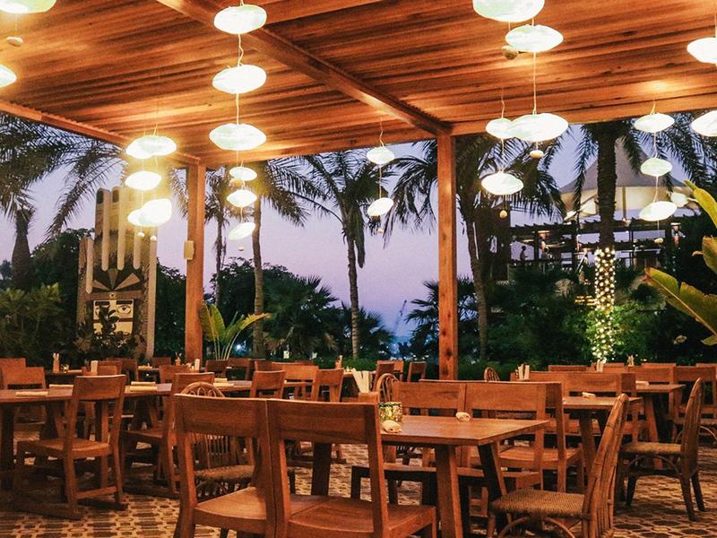 Offenes Restaurant mit Holzdecke, Hängelampen und Holztischen vor Palmen bei Dämmerung.