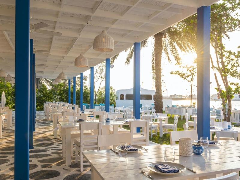 Freiluftrestaurant mit weißen Holztischen, blauen Säulen und Blick auf Wasser und Sonnenlicht.