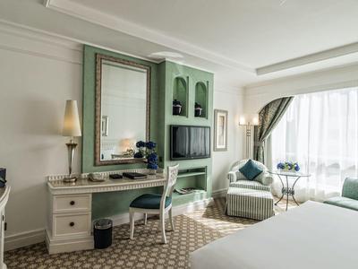 Helles Hotelzimmer mit großem Bett, Schreibtisch, Sessel, Fernseher und großem Fenster mit Vorhängen.