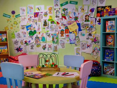 Bunter Kindertisch mit Spielzeug und Stiften, umgeben von Stühlen, Wand voller bunter Kinderzeichnungen.