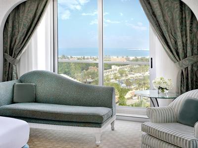 Wohnzimmer mit großen Fenstern, grüner Couch, Sessel, Vorhängen und Blick auf Stadt und Meer.