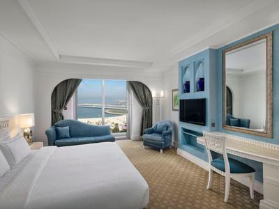 Helles Hotelzimmer mit großem Bett, blauen Möbeln, Meerblick und gemütlicher Sitzecke.