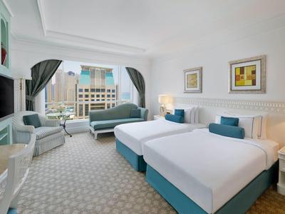 Helles Hotelzimmer mit zwei Einzelbetten, blauen Kissen, Sessel und großen Fenstern mit Vorhängen.
