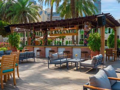 Outdoor-Bar mit Holzpergola, Sitzgruppen aus Holz und Metall, umgeben von Palmen und urbaner Kulisse.