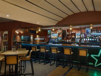 Innenraum einer Bar mit Holzmöbeln, Theke, Barhockern und grüner Sternbeleuchtung.