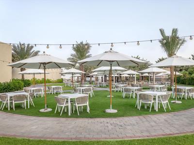Runder Außenbereich mit weißen Tischen, Stühlen und Sonnenschirmen auf Gras unter Lichterkette.