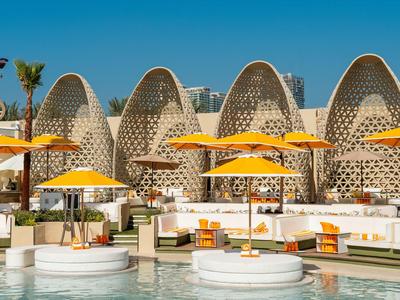 Moderner Hotelpool mit weißen Sitzinseln, orangen Sonnenschirmen und innovativer, bogenförmiger Architektur.
