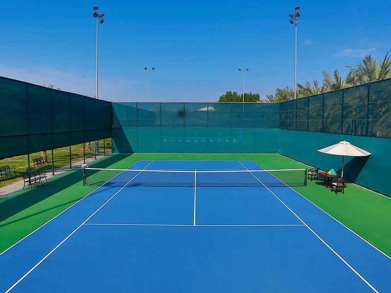 Leerer Tennisplatz mit blauem Belag, grünem Rand und umgeben von hohen Netzen bei sonnigem Himmel.