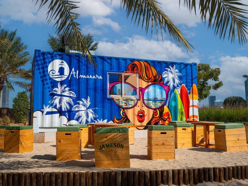 Bunte Strandkulisse mit Palmen, Sonnenbrille, Surfbrettern und Holzkisten vor blauem Himmel.