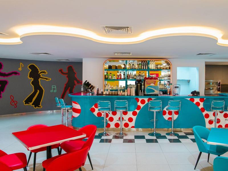 Moderner Raum mit farbigen Sitzmöbeln, Bar mit Getränkeauswahl und Wandkunst mit tanzenden Figuren.