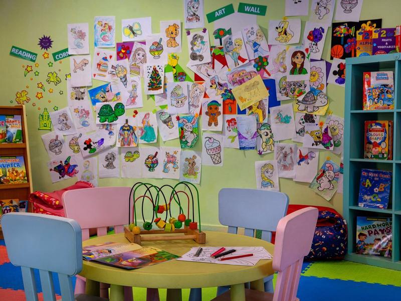 Bunter Kindertisch mit Spielzeug und Stiften, umgeben von Stühlen, Wand voller bunter Kinderzeichnungen.