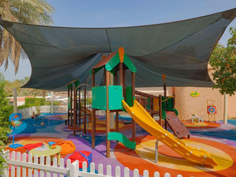 Bunter Spielplatz mit Rutsche, Klettergerüst, Sonnenschutz und weißem Zaun unter Bäumen.