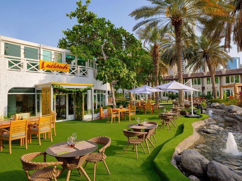 Außenbereich mit Tischen und Stühlen, grüner Rasen, Palmen und Wasserspiel neben weißem Gebäude.