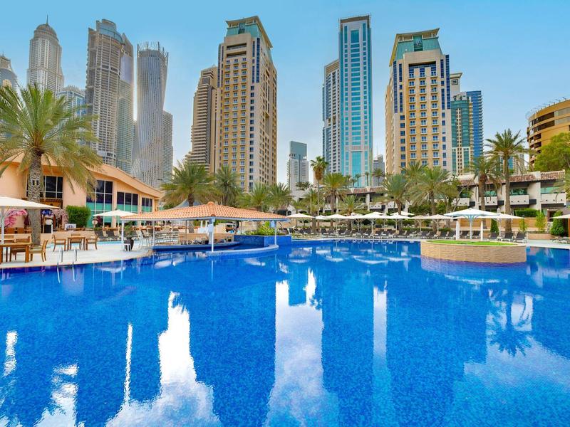 Großer blauer Pool vor modernen Wolkenkratzern und Palmen bei klarem Himmel.