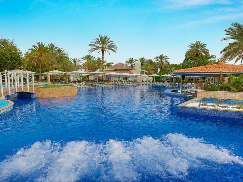 Großer blauer Pool mit Palmen, Sonnenschirmen und Liegestühlen bei klarem Himmel.