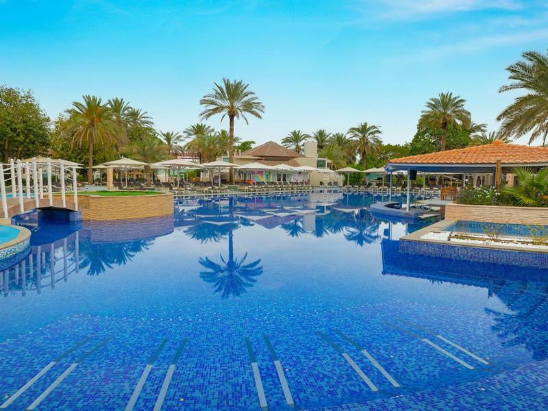 Großes blaues Schwimmbecken mit Palmen, Sonnenschirmen und Sitzbereichen bei klarem Himmel.