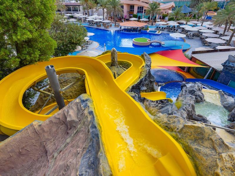 Eine leuchtend gelbe Wasserrutsche windet sich um Felsen in einem Poolbereich bei Sonnenschein.