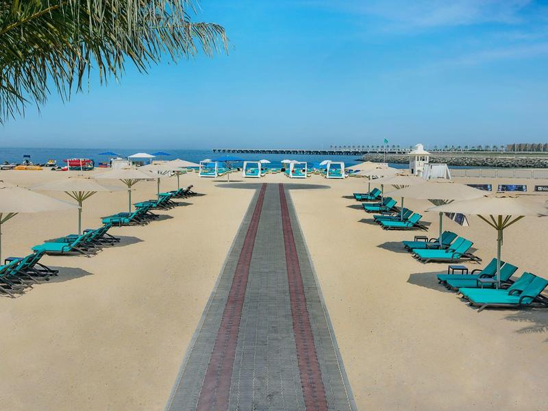 Strand mit Liegen, Sonnenschirmen und klarem blauen Himmel über ruhigem Meer.