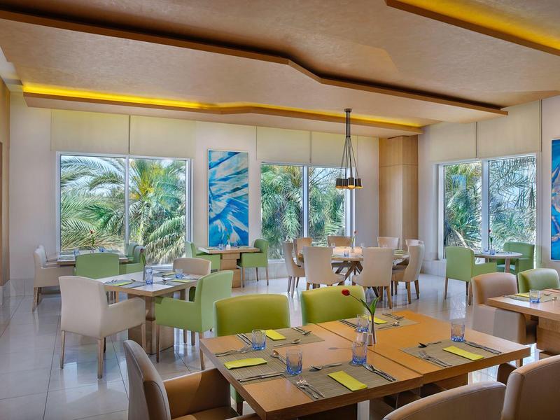 Helles Restaurant mit großen Fenstern, grünen Pflanzen draußen, weißen Tischen und beigen Stühlen.