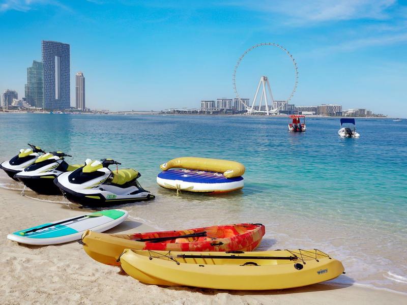 Mehrere bunte Kajaks, Jet-Skis und eine aufblasbare Plattform stehen am Sandstrand vor klarem blauem Meer.