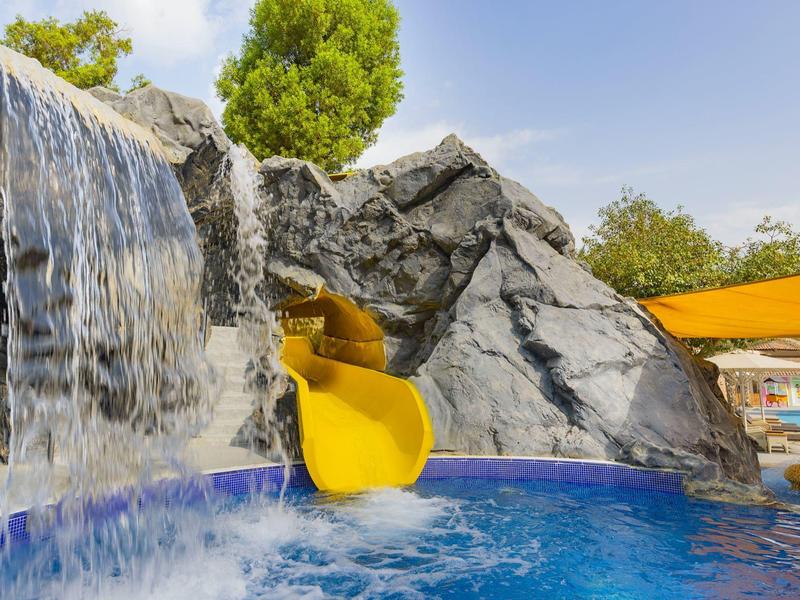 Wasserfall und gelbe Rutsche neben künstlichen grauen Felsen über blauem Pool.