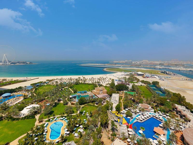 Blick auf Küstenlandschaft mit Sandstrand, Meer, Hotels, Pools und grüner Vegetation bei blauem Himmel.
