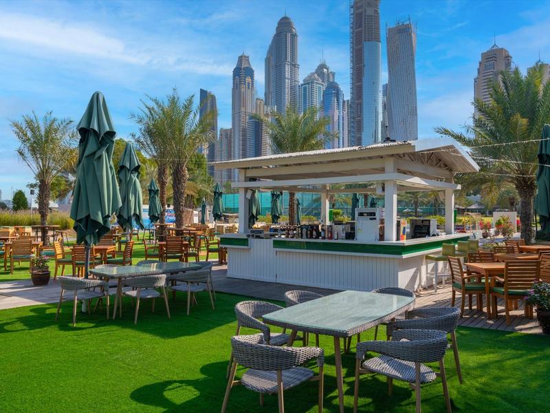 Grüne Terrasse mit Tischen, Stühlen und Sonnenschirmen vor Wolkenkratzern bei blauem Himmel.