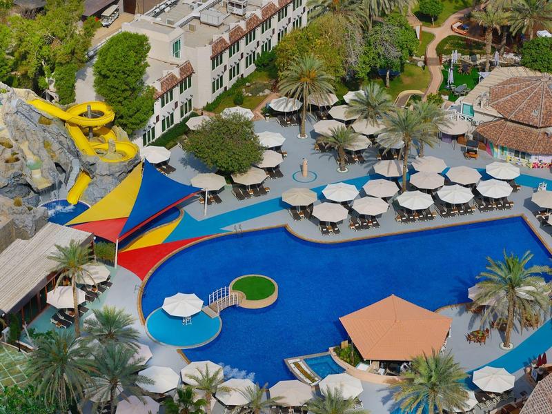 Großer blauer Swimmingpool mit Sonnenschirmen, Liegestühlen, Palmen und Wasserrutschen in Resortanlage.