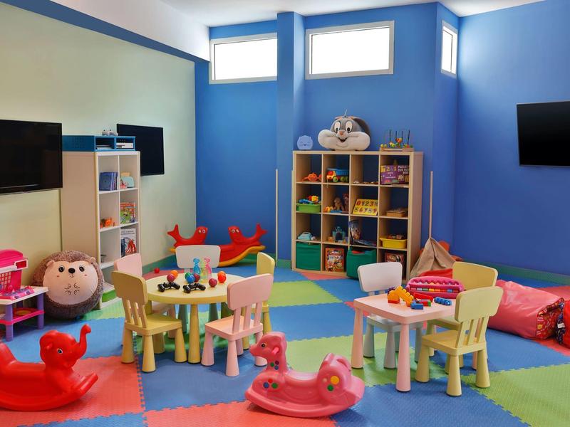 Bunter Kinderraum mit Spielzeug, kleinen Tischen, Stühlen und weichen Sitzkissen an blauen Wänden.