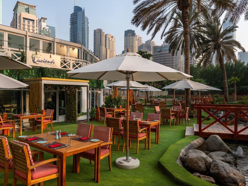 Außenterrasse mit Holztischen und weißen Sonnenschirmen neben einem Teich und Palmen in einer Stadt.