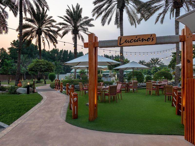 Außenbereich eines Restaurants mit Tischen unter weißen Sonnenschirmen und Palmen im Hintergrund.