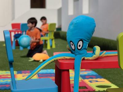 Bunter Spielbereich mit bunten Stühlen, Teppichen und einem blauen Tintenfisch-Charakter.