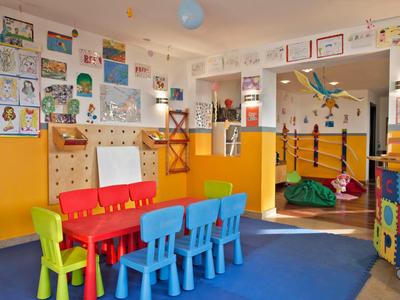 Bunter Kindergartenraum mit kleinen Stühlen, Maltisch, Kunstwerken an den Wänden und weichem Boden.