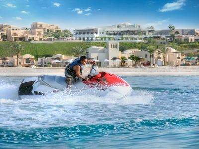 Mann fährt Jetski auf blauem Wasser vor Strand mit Gebäuden und blauem Himmel.