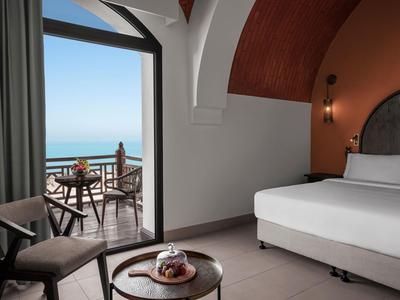 Modernes Hotelzimmer mit Bett, Stuhl, Balkon und Blick auf das Meer bei sonnigem Wetter.