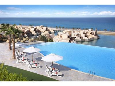 Infinity-Pool mit weißen Liegestühlen und Sonnenschirmen vor Meer und modernen Bungalows am Wasser.